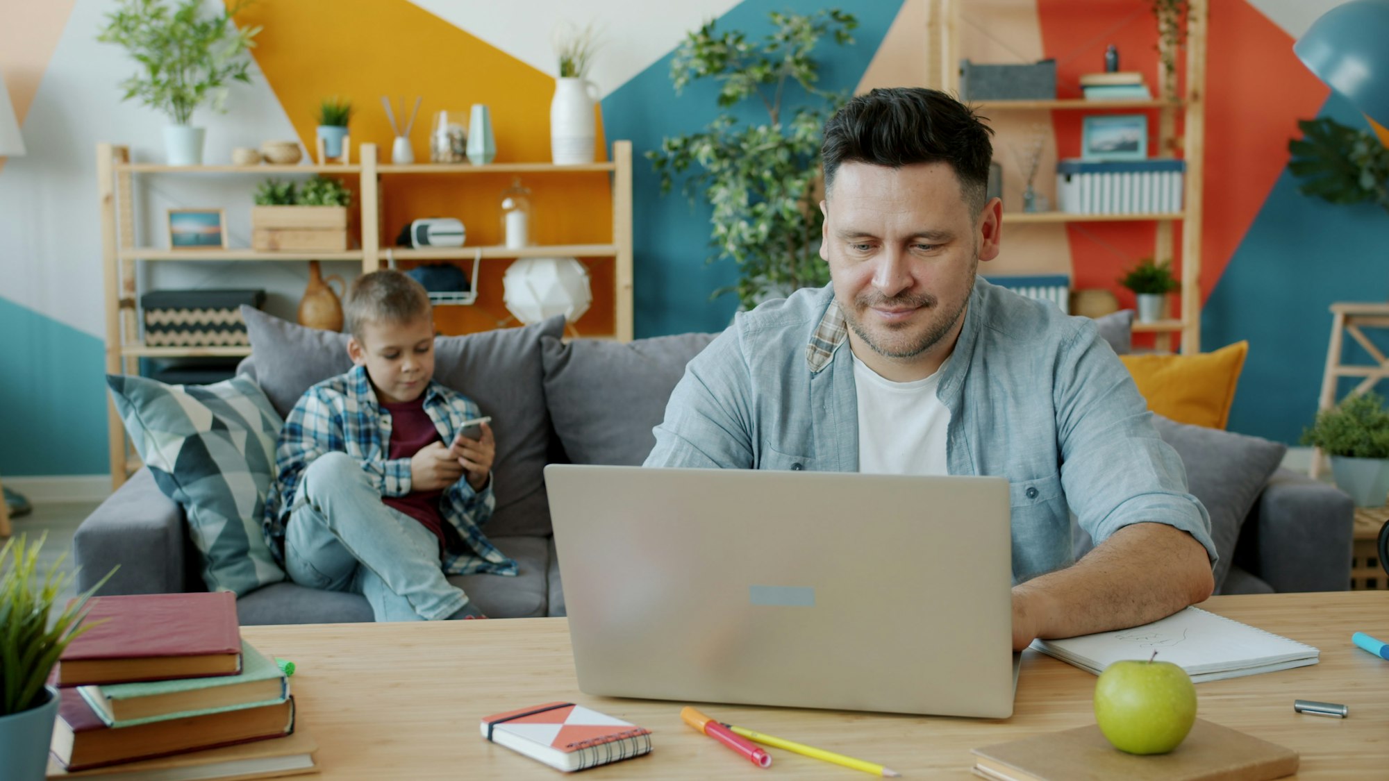Father works on laptop while son plays on phone beside him