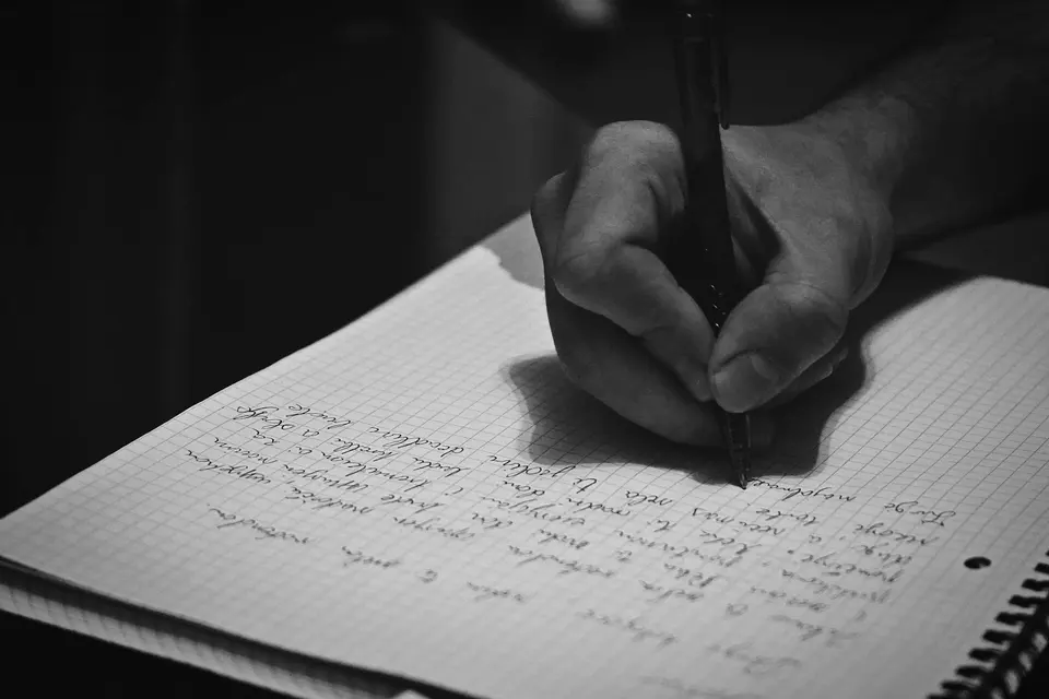 A black-and-white photograph of a hand writing in a notebook