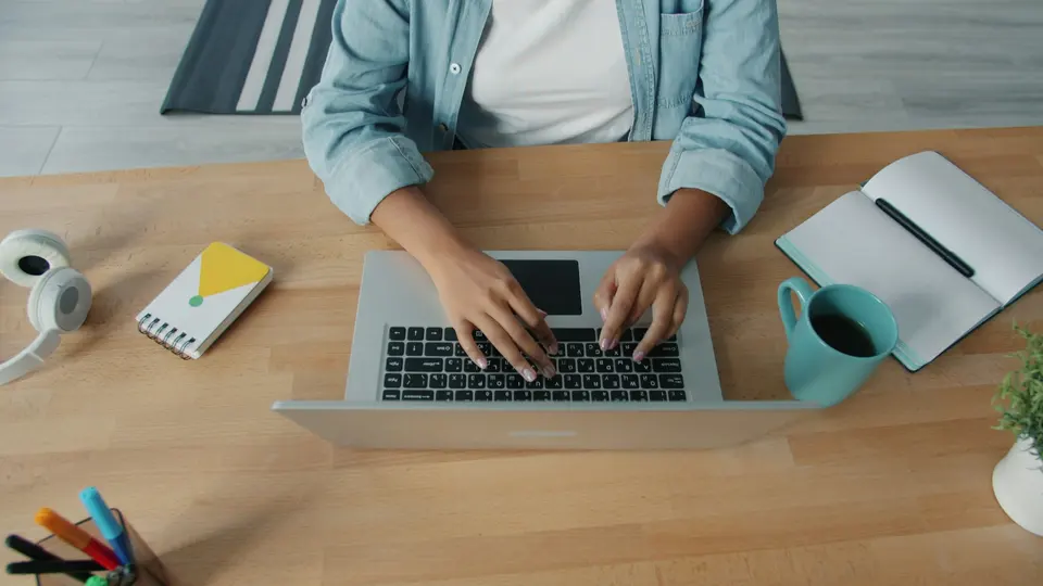 A person typing on a laptop beside notebooks and mugs