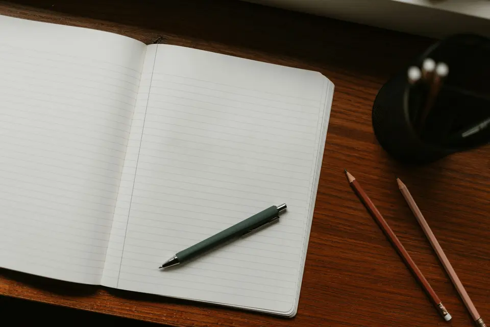 An open blank notebook with a pen and pencils on a wooden desk